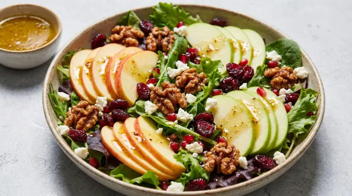A vibrant green salad topped with red apple slices, cranberries, and candied walnuts in a large serving bowl.